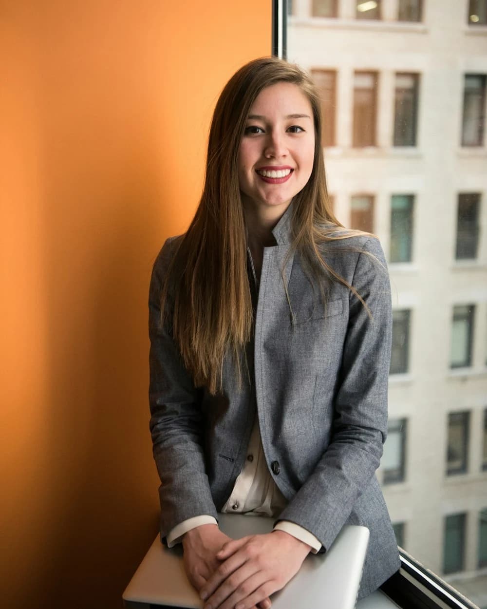 Smiling executive assistant in a blazer holding a laptop by an office window