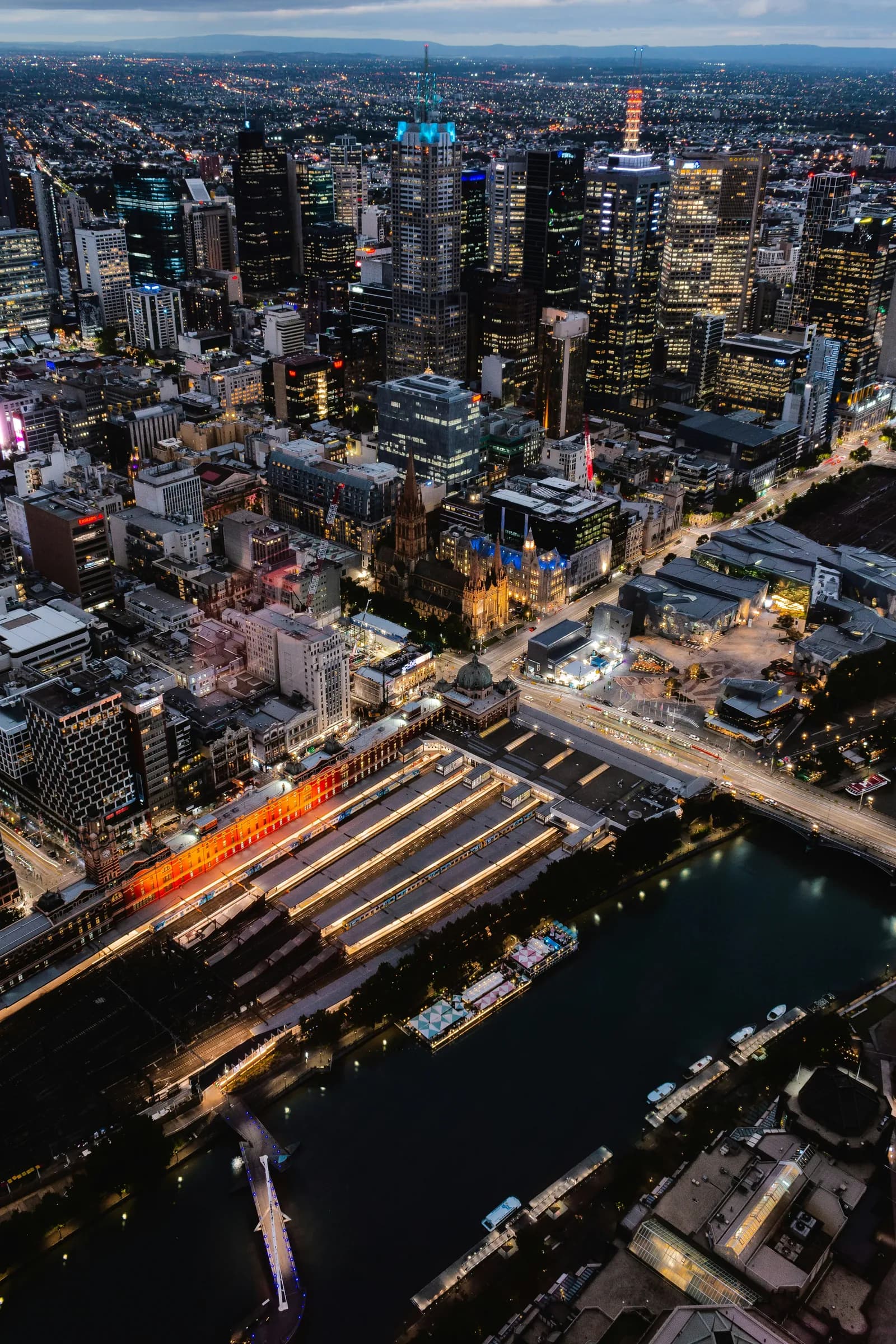 Melbourne's city centre from the air at dusk — Flinders Street Station, Federation Square and the Yarra, where ME CABS hotel transfers finish